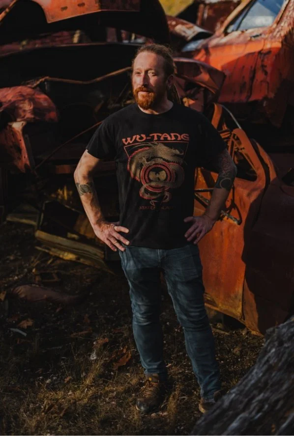 A man with a red beard and tattoos on his arms stands outdoors in front of a pile of rusted, damaged, and burned cars, wearing a black T-shirt with a graphic design and the words 'WU-TANG' on it, and jeans.