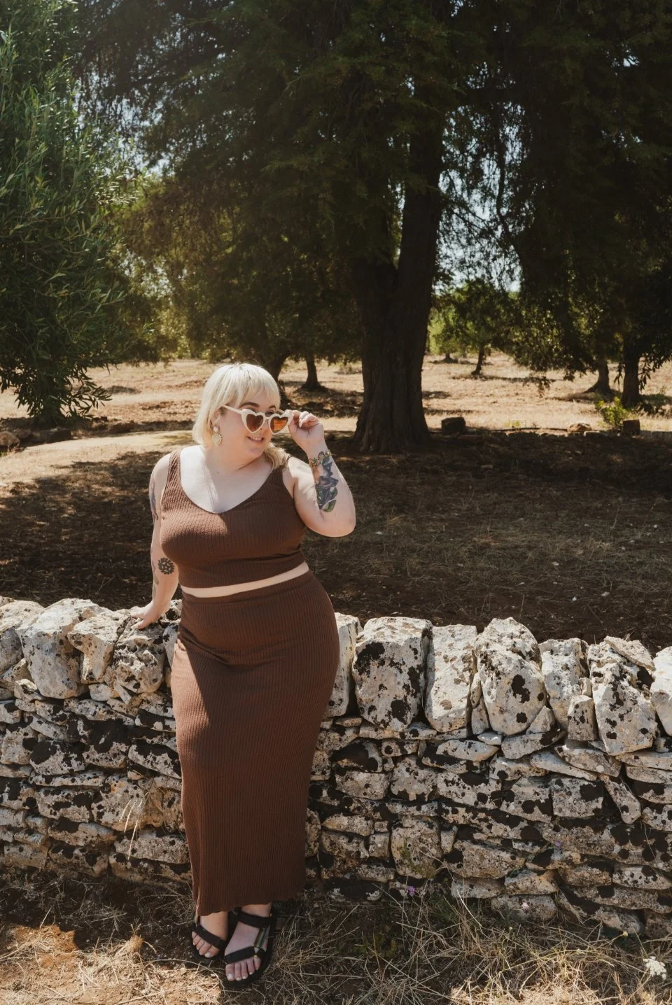 A woman with blonde hair and tattoos wearing heart-shaped sunglasses and a brown sleeveless dress standing by a stone wall in Tuscany, in a sunny outdoor setting with trees in the background.