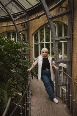 Woman with short white hair standing in an industrial-style glass and metal structure surrounded by green plants.