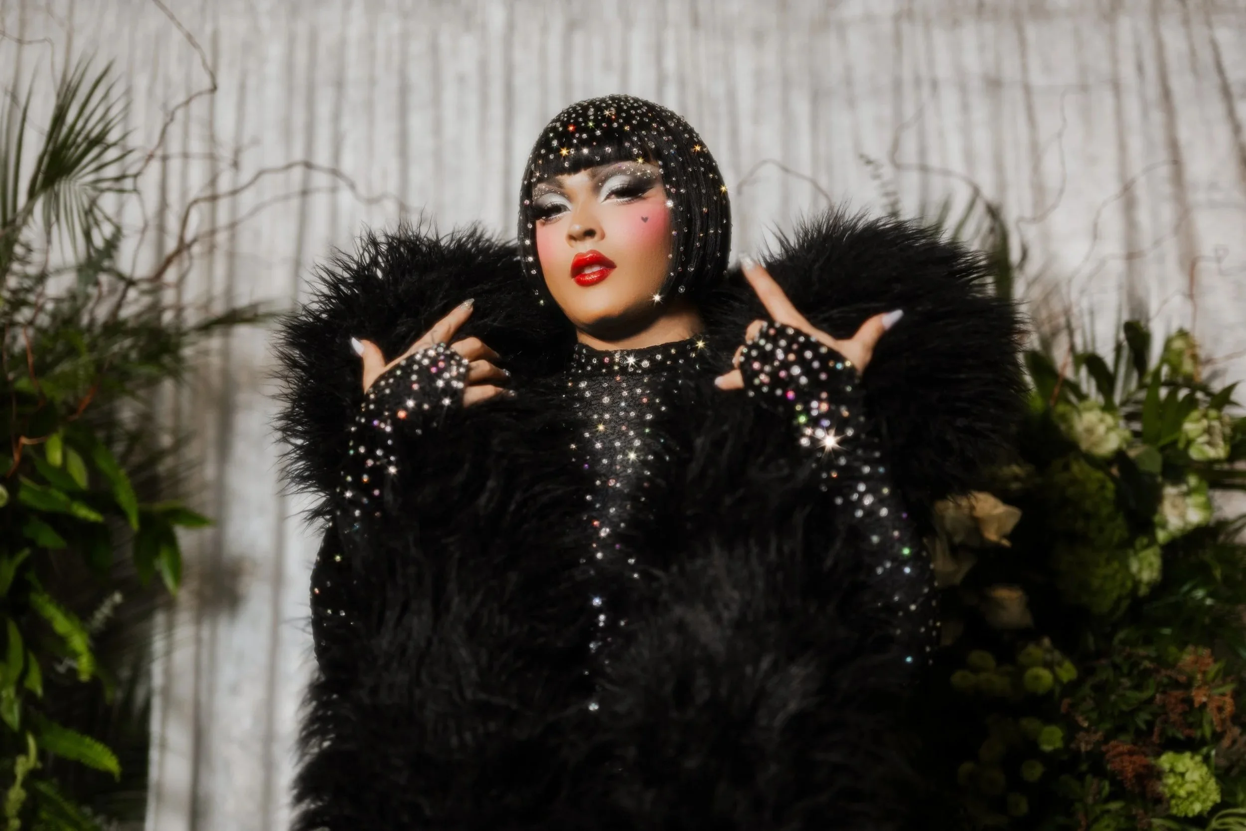 A woman with short black hair styled in a bob, adorned with rhinestones, wearing heavy makeup with silver eyeshadow and red lipstick, dressed in a black, sparkling, fluffy fur coat, posing with her hands near her face against a background of plants and a textured wall.