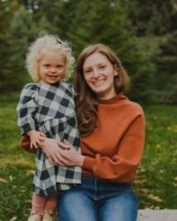 A woman with long hair wearing an orange sweater holding a young girl with curly blonde hair in a plaid dress outdoors with trees in the background.