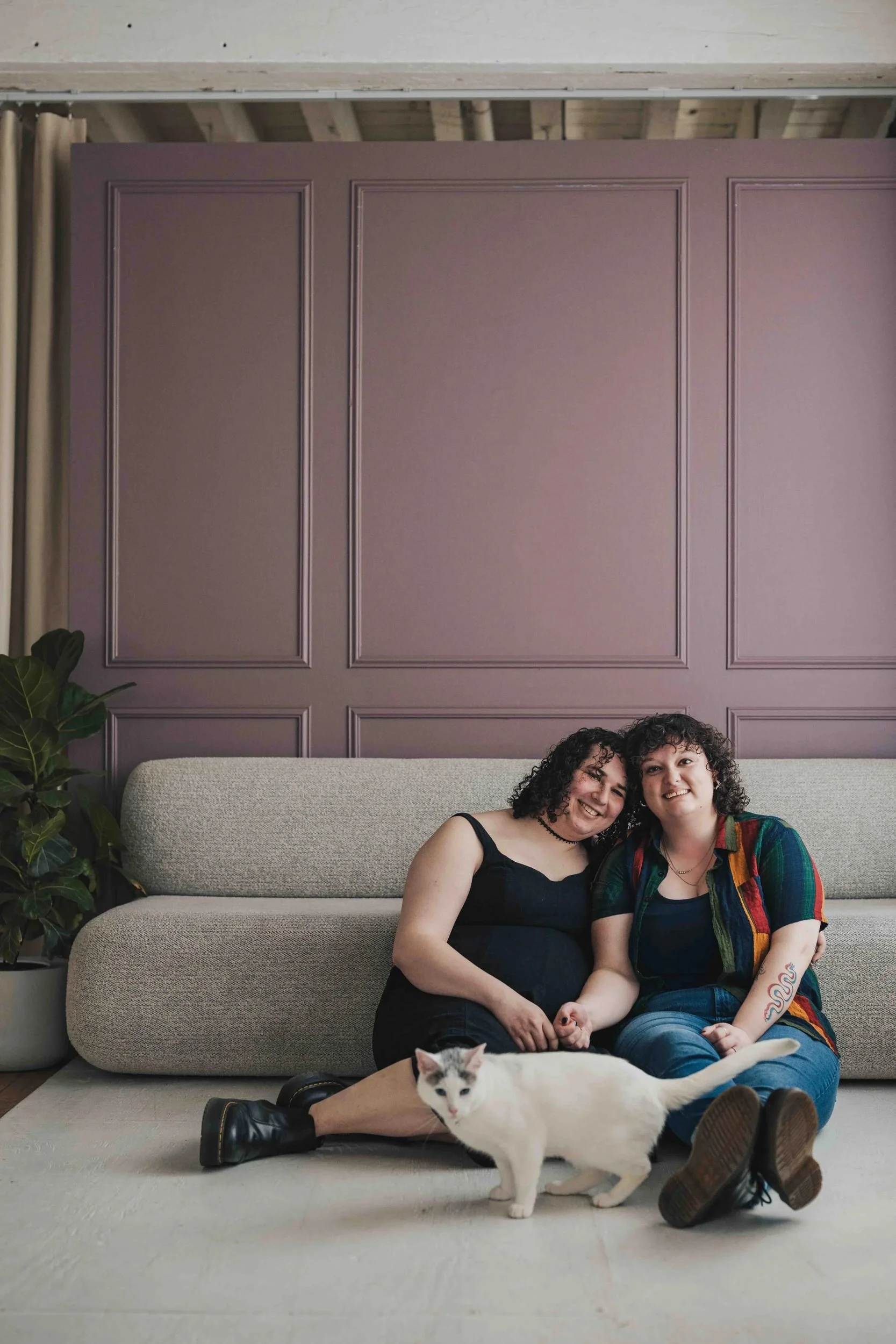 Two women with curly hair sitting on the floor in front of a beige couch with purple wall panels behind them. A white cat with black markings lies on the floor near their feet, and a large potted plant is on the left side of the image.