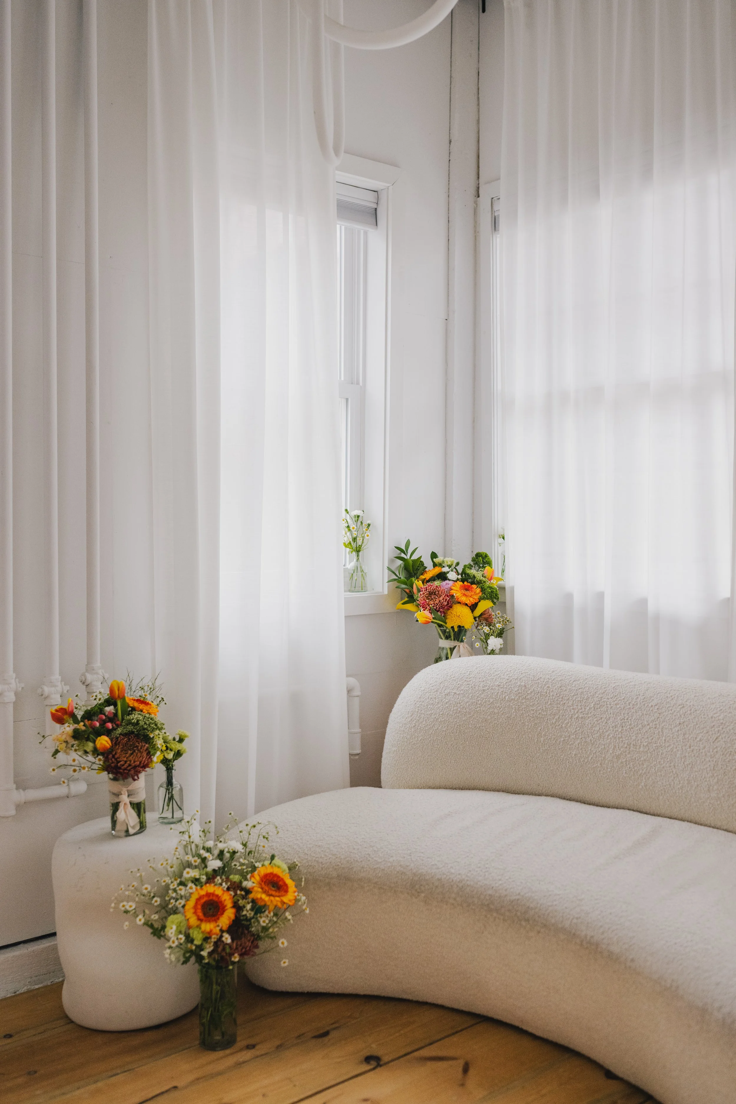 A cozy room with white sheer curtains; a floral arrangement with orange, yellow, and pink flowers in a glass vase; a white upholstered armchair with rounded edges; and a white side table with a smaller bouquet of flowers, all on a wooden floor.