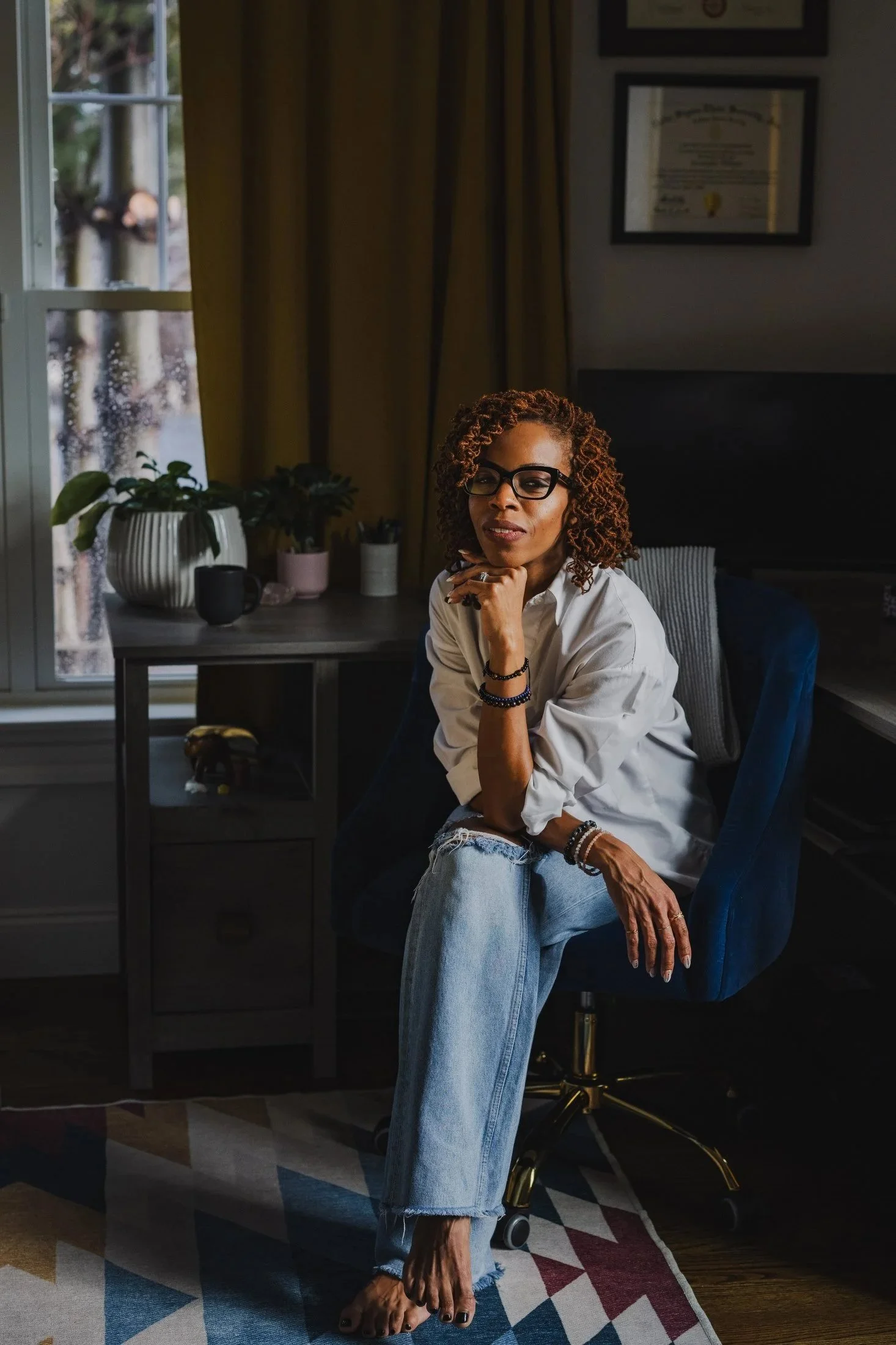 A woman with curly hair, glasses, and a white shirt sitting on a chair in a room with books and plants, holding her chin, and smiling slightly.