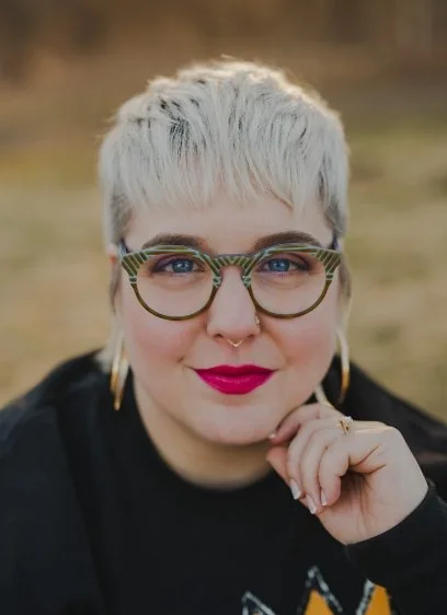 Close-up of a woman with short platinum blonde hair, colorful glasses, gold earrings, and pink lipstick, outdoors.
