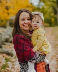 A woman holding a baby girl in an outdoor park with autumn foliage.
