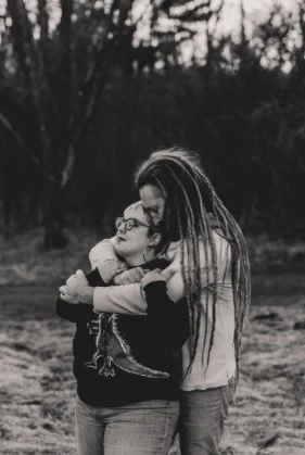 Two people hugging outdoors, one with dreadlocks and the other with glasses, in black and white.