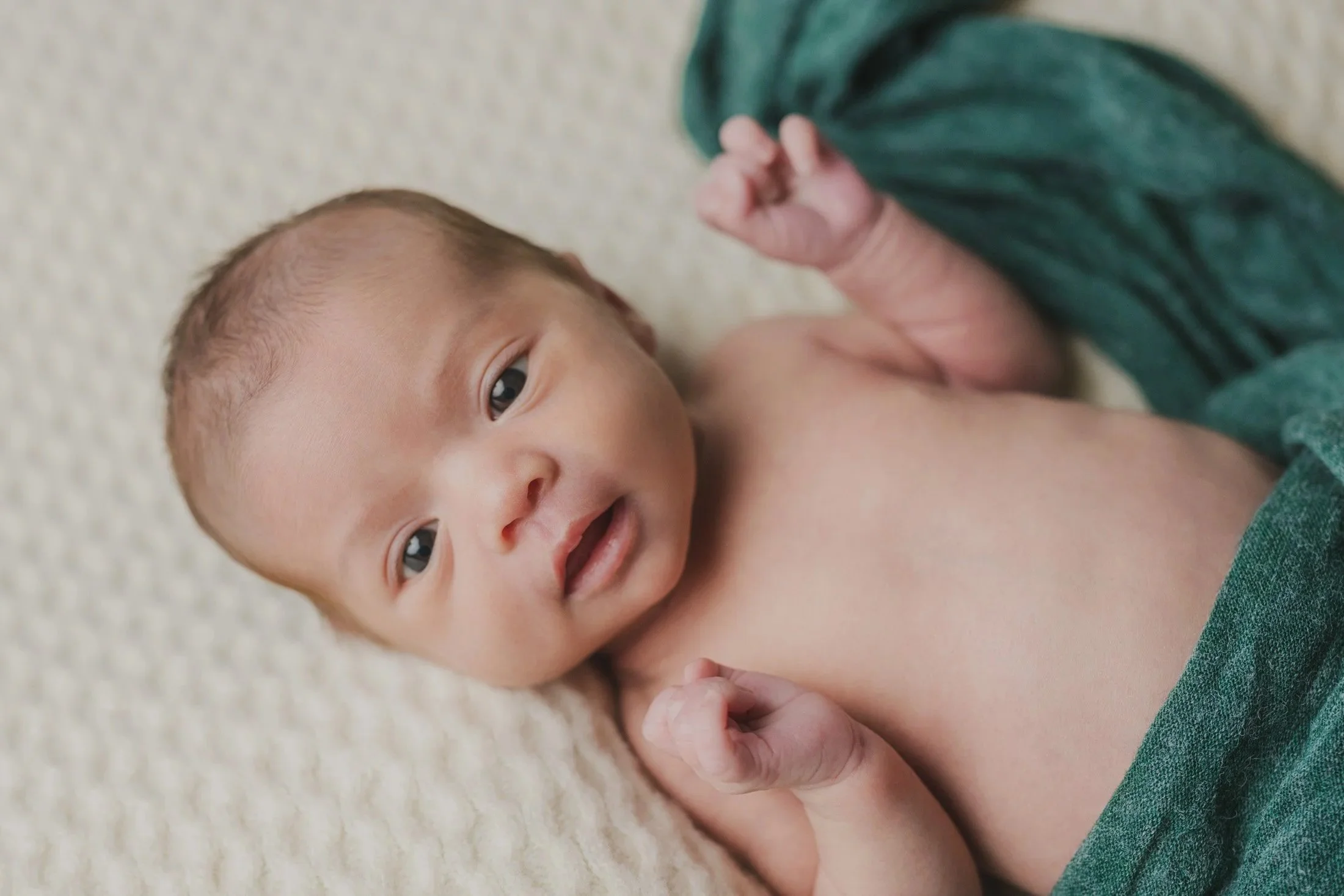 Close-up of a baby laying on a soft light-colored surface, looking up with curious eyes, wearing a green outfit.