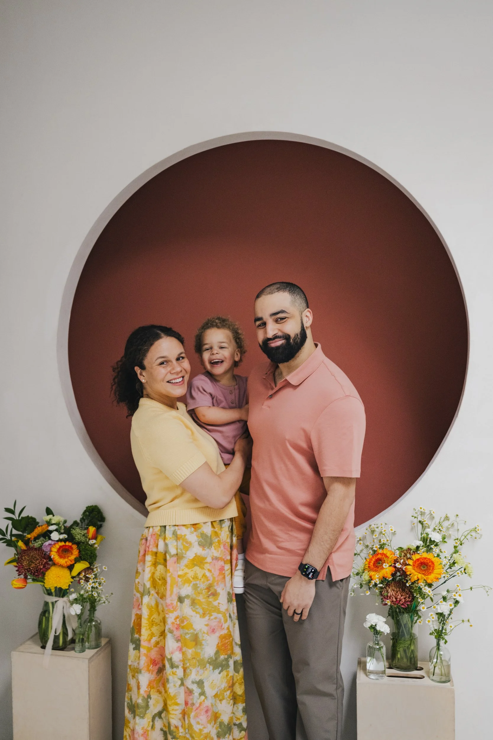 A family of three posing in front of a circular backdrop with a brown interior. The mother, wearing a yellow top and floral skirt, is holding a young girl with curly hair and a purple dress. The father, with a beard and wearing a pink shirt with a smartwatch, stands beside them. There are colorful flower arrangements on either side of them.
