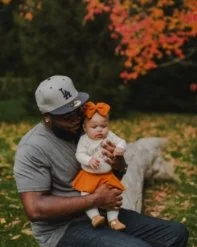 Man holding a baby girl outdoors in fall foliage, both wearing casual clothes and the girl has an orange bow in her hair.