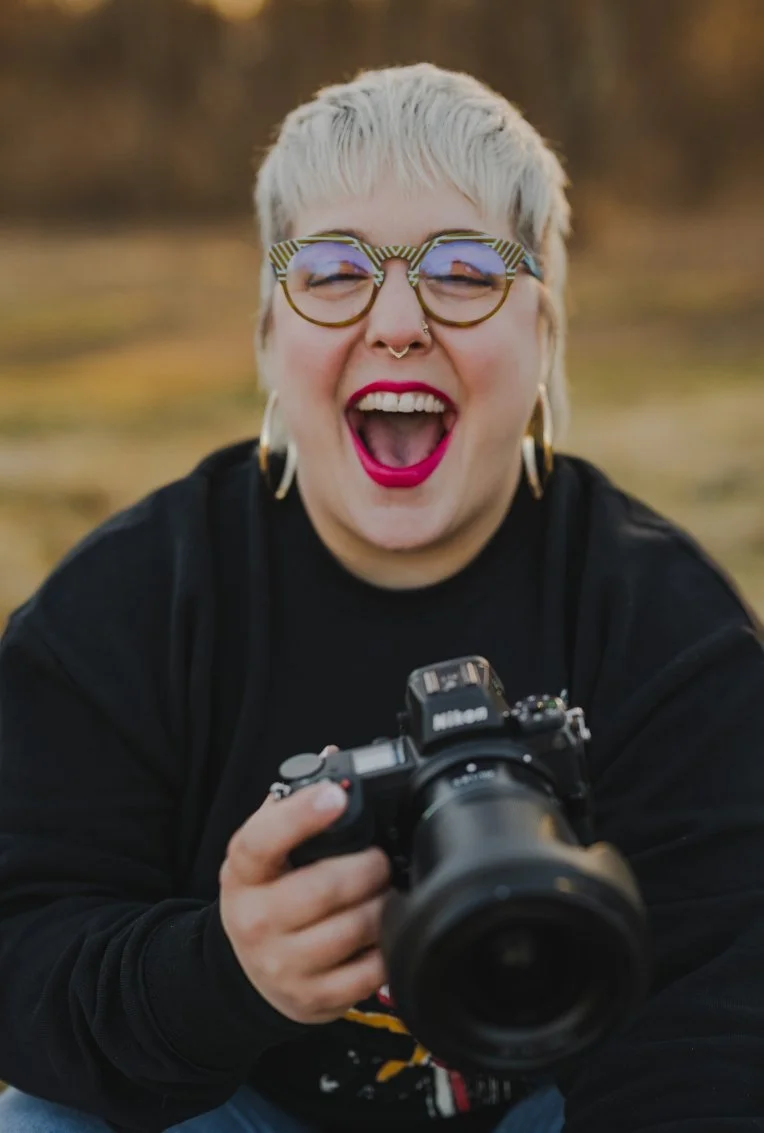 A person with short, bleached blonde hair and glasses with striped frames, wearing large hoop earrings and bright pink lipstick, laughs joyfully while holding a camera outdoors.