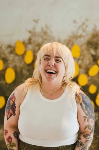 A young woman with blonde hair and tattoos smiling and laughing in front of a background of yellow flowers and greenery.