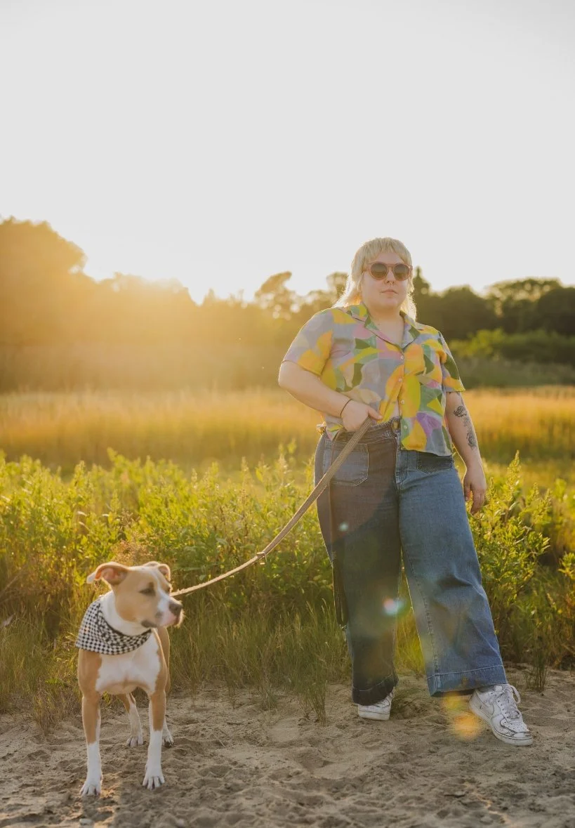 A woman with tattoos, wearing sunglasses, a colorful short-sleeve shirt, and baggy jeans, holding a leash attached to a brown and white dog with a black and white checkered bandana around its neck. They are outdoors in a grassy area during sunset.