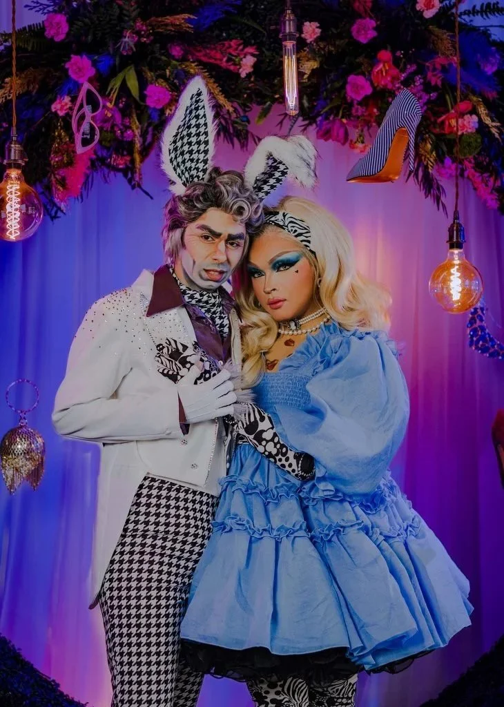 Two drag artists dressed in elaborate, colorful costumes with animal ears and makeup, standing under a floral and decorative backdrop with hanging light bulbs and high-heeled shoes.