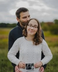 A man and woman standing outdoors in a field, with the woman holding an ultrasound photo, both smiling.