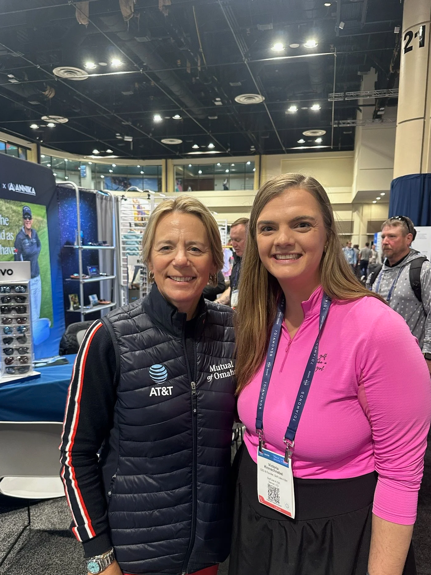 Two women standing together at a conference or event with booths and other attendees in the background. One woman is wearing a black jacket with a logo on it, and the other woman is wearing a pink top with a name tag. They are smiling at the camera.