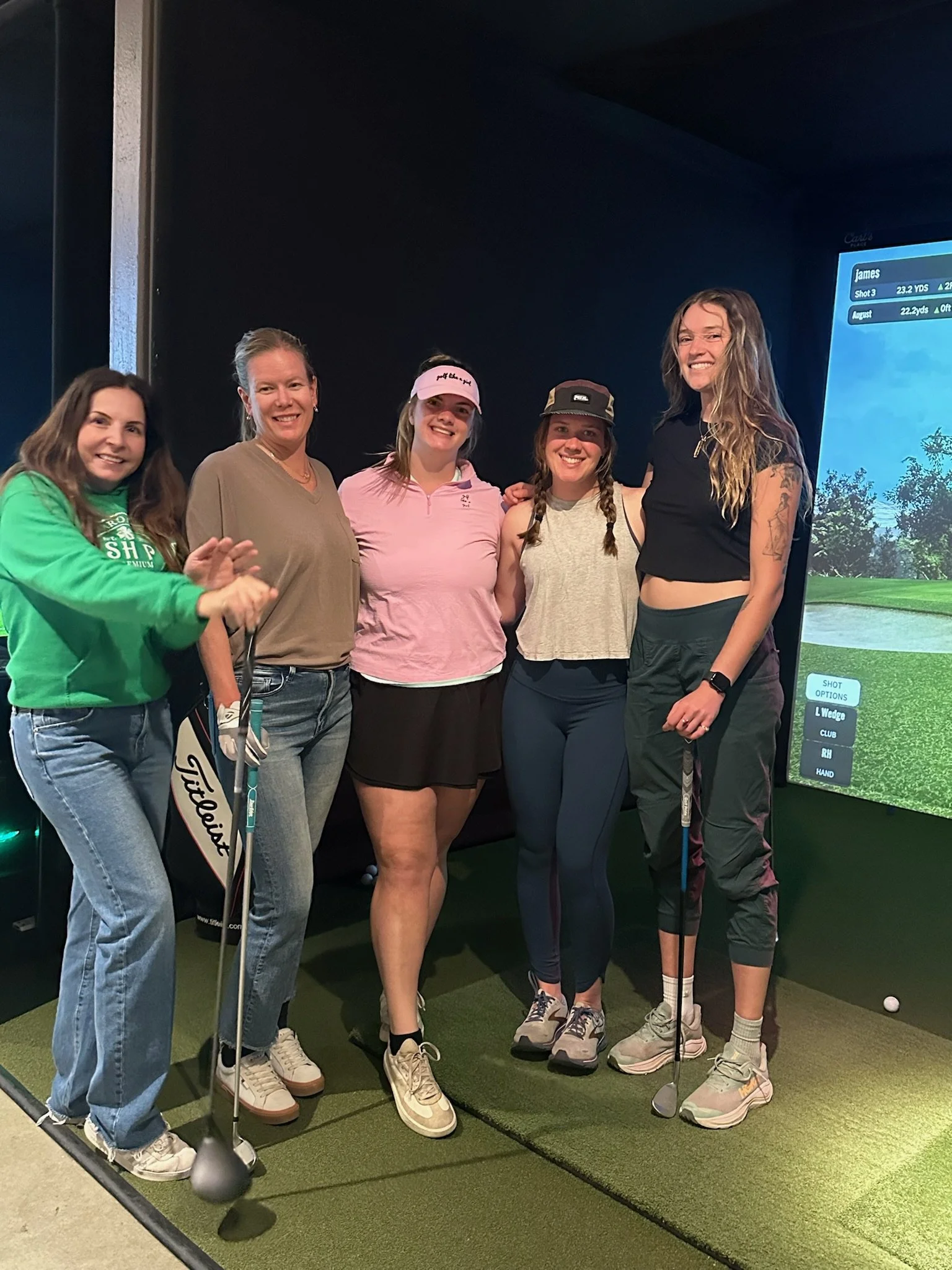 Five women standing together at an indoor golf simulator, holding golf clubs, smiling for the photo.