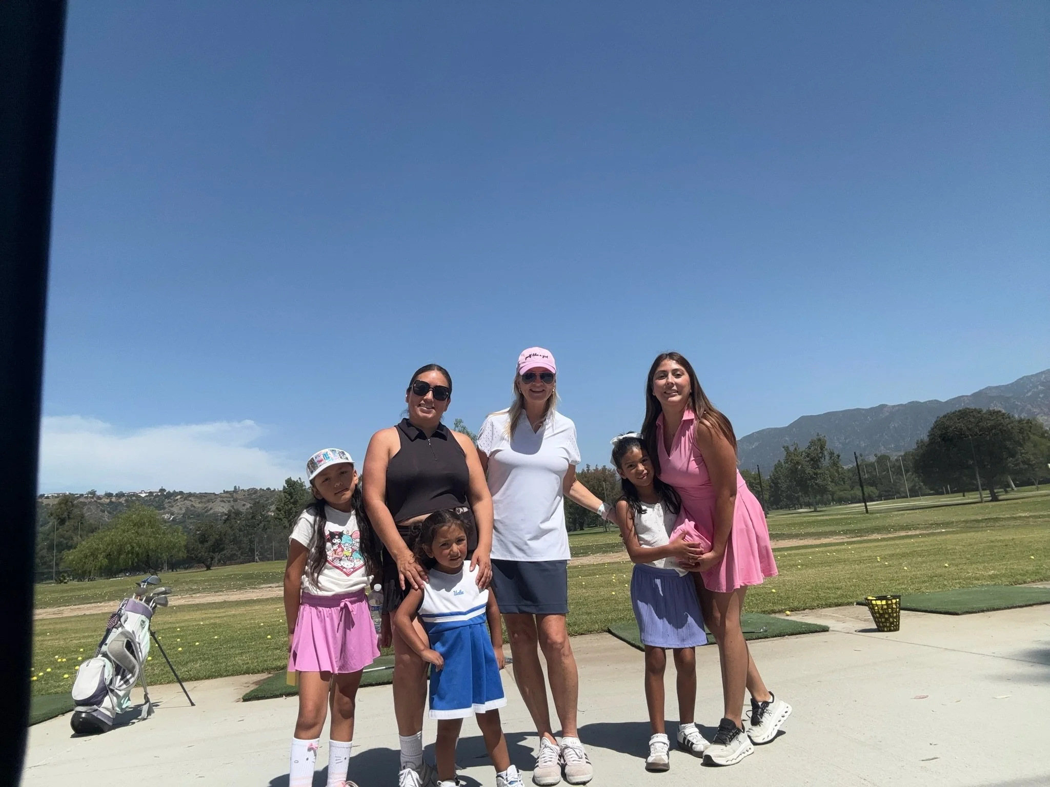 Group of young girls and women posing on a golf course with mountains in the background, sunny weather, some wearing sunglasses and hats.