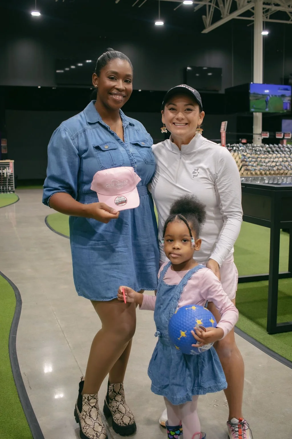 Two women and a young girl at a miniature golf course. The woman on the left is holding a pink cap, the woman on the right is smiling, and the girl is holding a blue ball with yellow stars, wearing a denim dress over pink shirt and white tights.