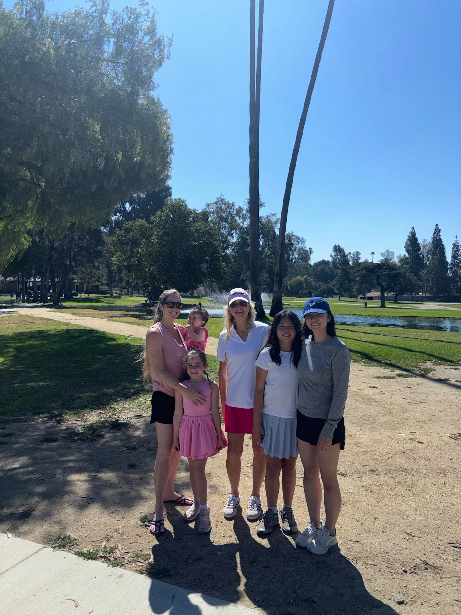 Group of six women and girls standing outside on a sunny day near a pond, with trees and a water fountain in the background.