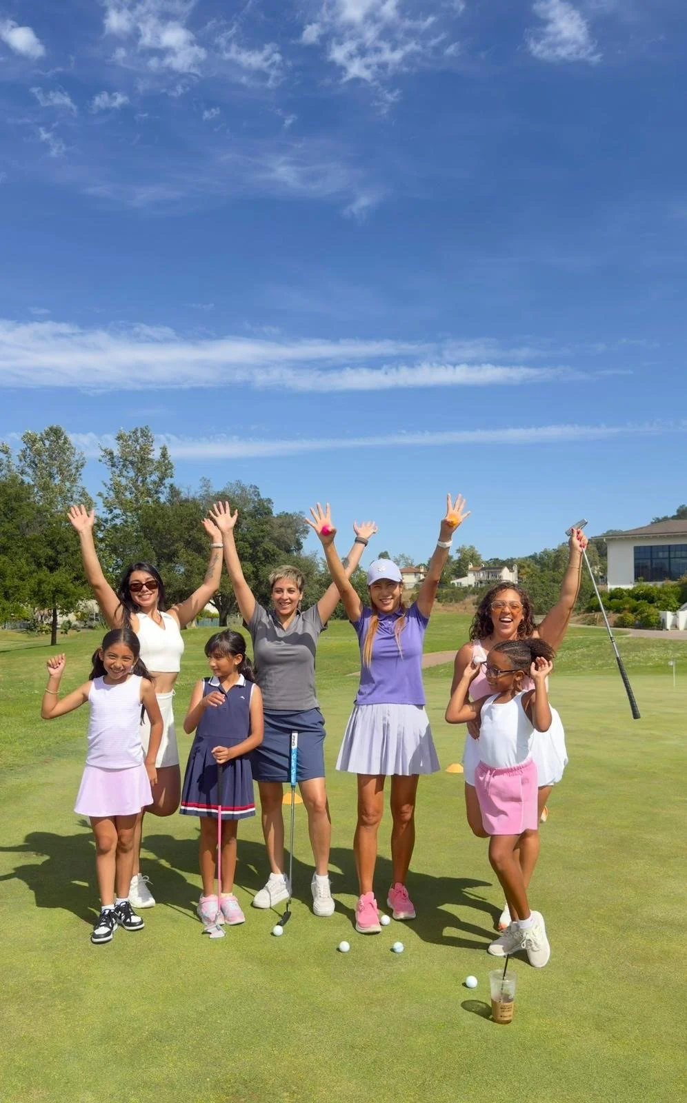 Group of women and girls celebrating on a golf course, raising their arms in joy, with golf balls and a drink on the grass, under a blue sky with clouds.