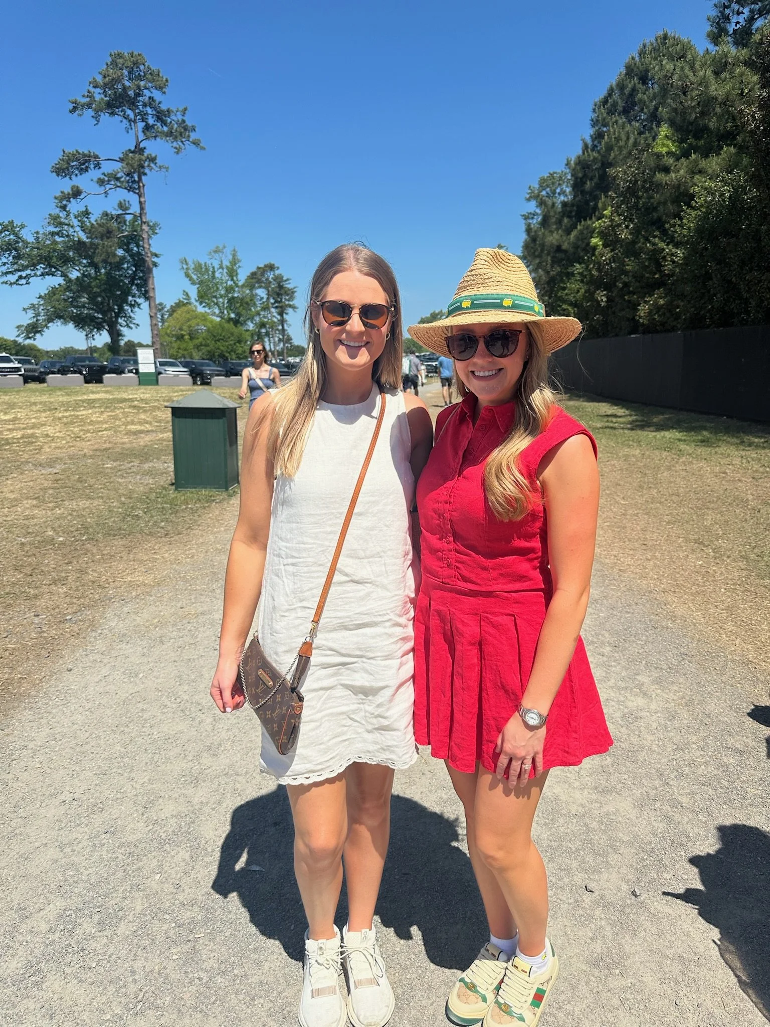 Two women standing outdoors on a sunny day, smiling at the camera. One is wearing a white dress, sunglasses, and white sneakers, and the other is in a red dress, sunglasses, a straw hat, and colorful sneakers. There are trees, parked cars, and other 