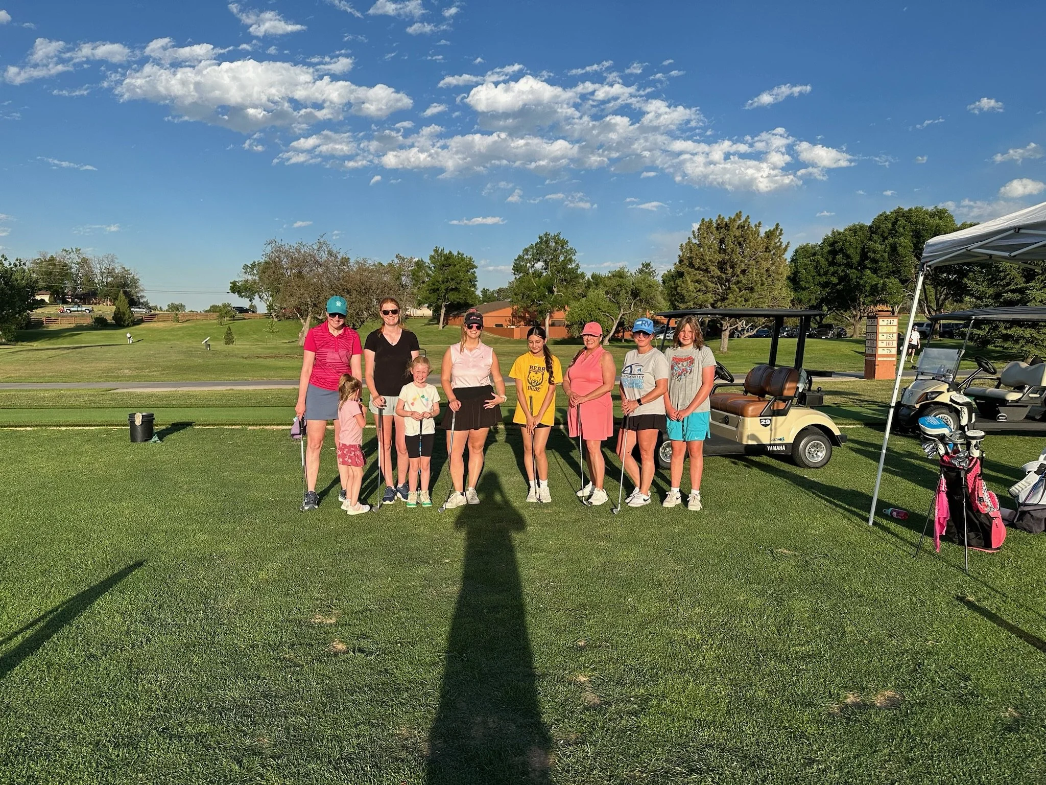 A group of ten people, mostly children, standing on a golf course with two golf carts nearby. The background features a bright blue sky with scattered white clouds and green trees.