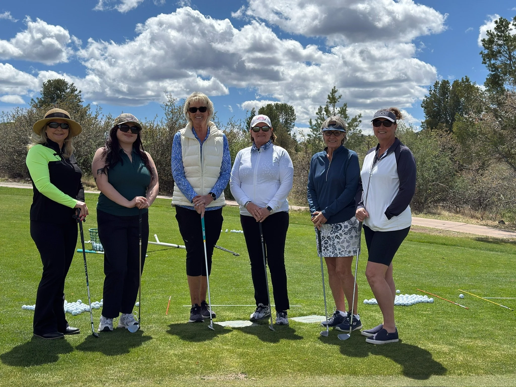 A group of six women standing on a grassy golf course, holding golf clubs, with golf balls on the ground behind them, under a partly cloudy sky with trees in the background.