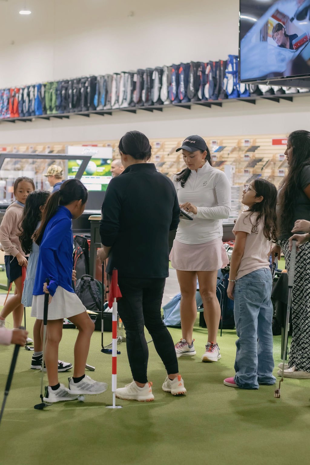 A group of young girls and women on an indoor golf practice range, with golf clubs, a small flag, and tables with golf gear around.