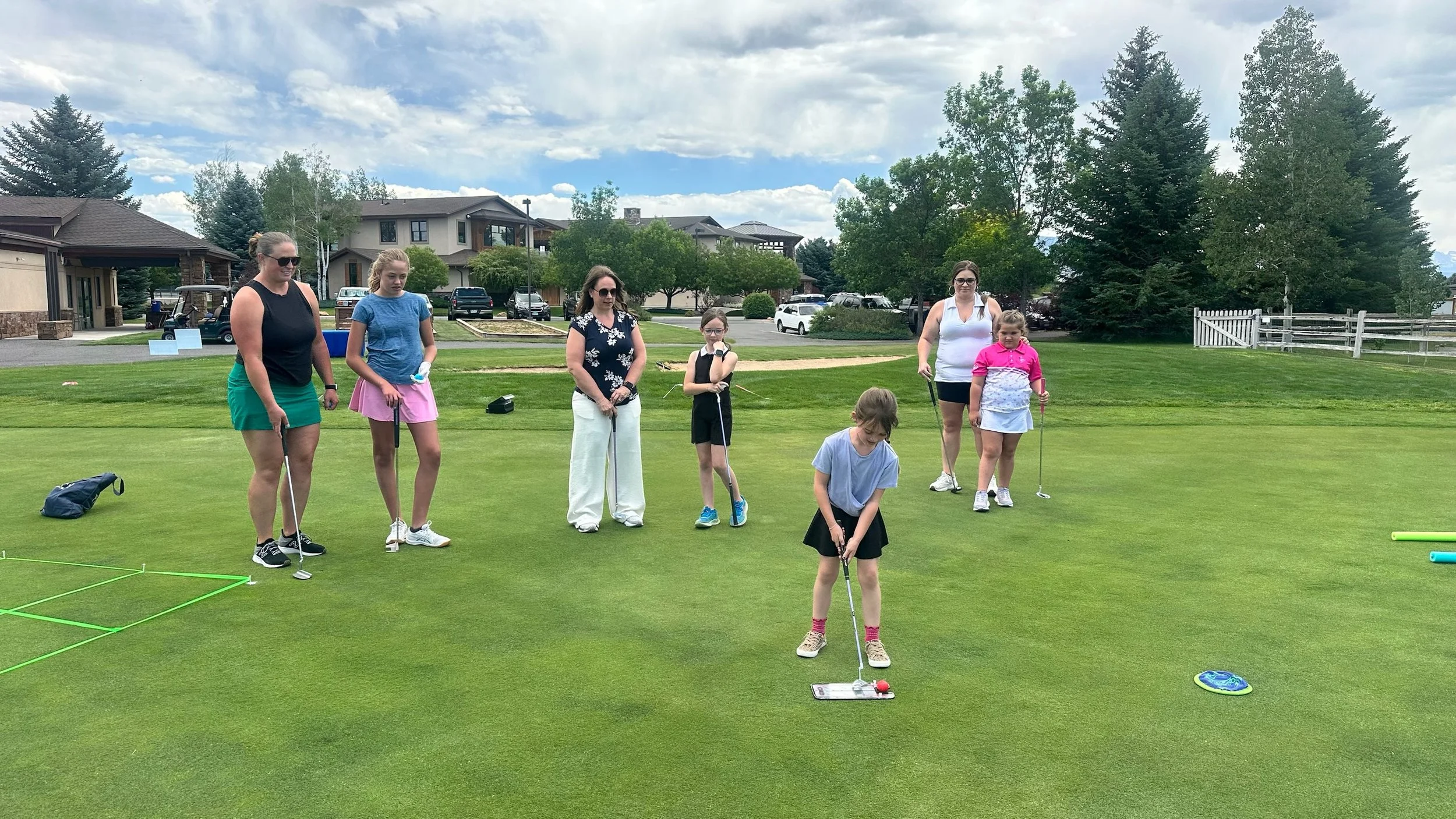A group of seven people, including children and adults, are standing on a golf putting green, with some using putters to practice putting. The scene is outdoors on a clear day with scattered clouds, in a residential area with houses, trees, and parke
