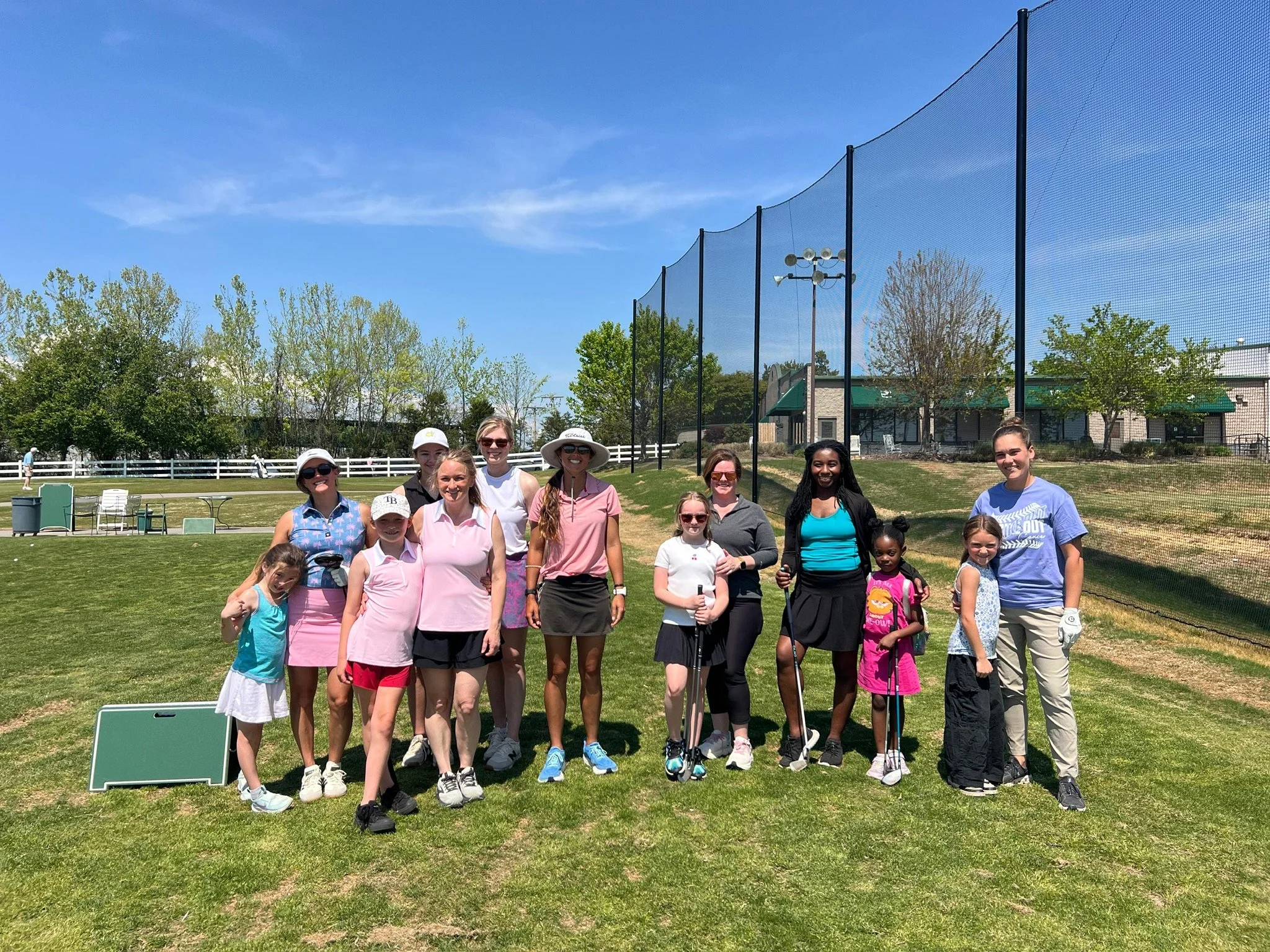 A group of women and children posing on a grassy field near a fenced sports area on a sunny day, with trees, a building, and blue sky in the background.
