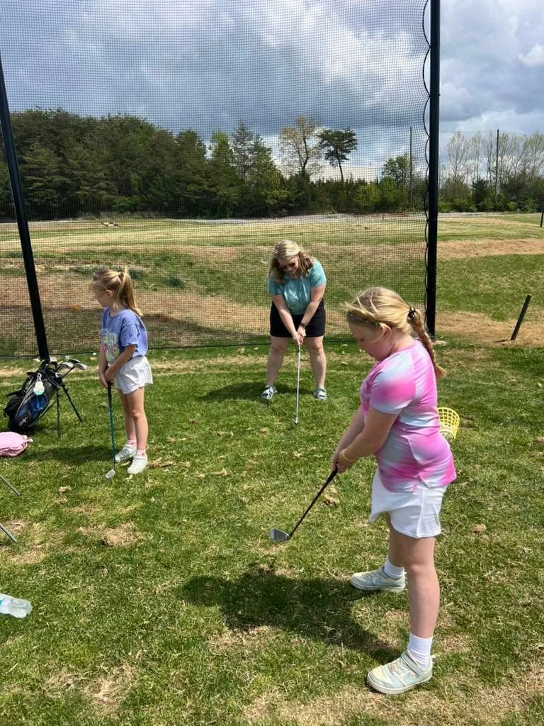 Three young girls and a woman playing mini golf outdoors on a sunny day, with a netted enclosure and trees in the background.