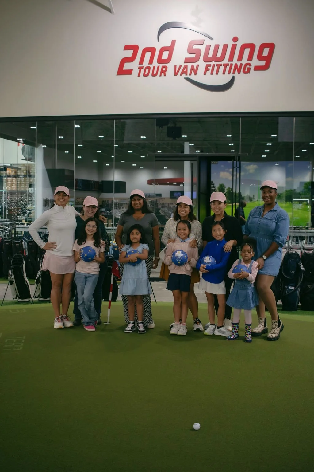 Group of women and children posing on a golf practice putting green at a golf facility called '2nd Swing Tour Van Fitting.' The group includes young girls and adult women, some holding blue golf balls, wearing casual clothing and pink caps. They are 
