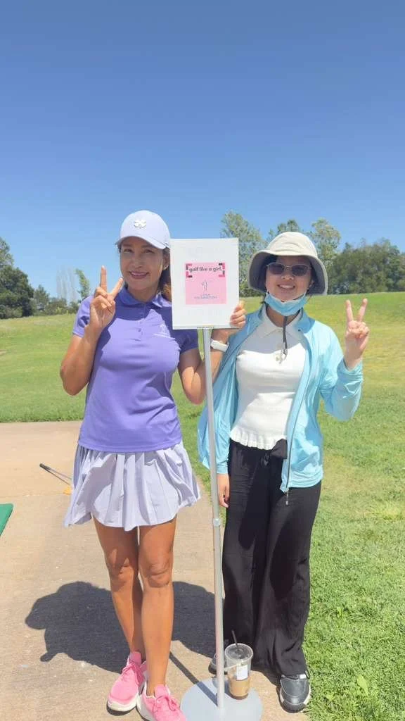 Two women standing outdoors on a golf course, one holding a sign that says 'Feel like a girl,' and both making peace signs, under clear blue skies and green trees.