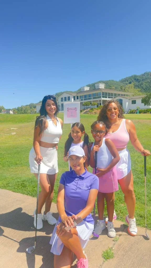 A group of five women and girls outdoors on a sunny day, dressed in golf attire, holding golf clubs, with a green field and a building in the background.