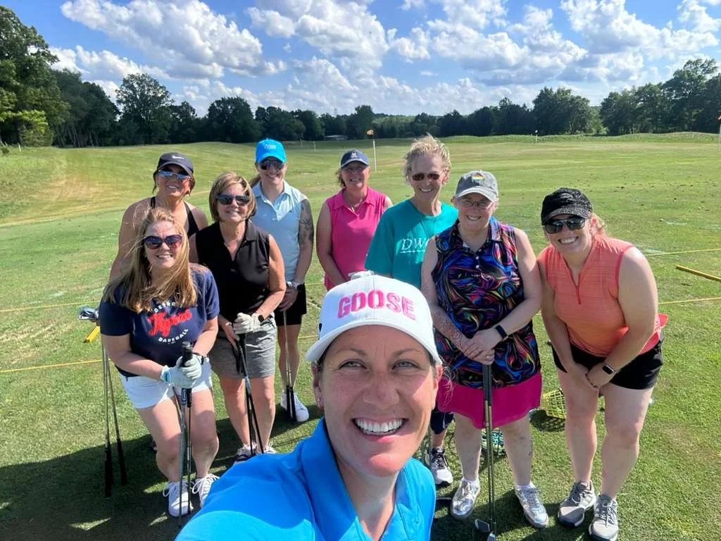 A group of ten women on a golf course, smiling and posing for a selfie. They are wearing casual golf attire and have golf clubs.