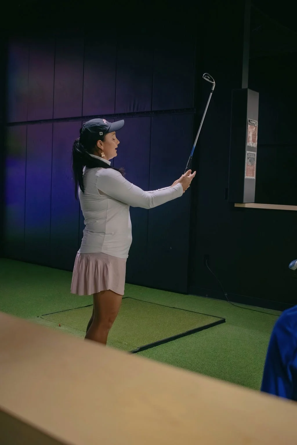 A woman wearing a golf cap, white top, and pink skirt holding a golf club indoors, preparing to take a shot on a green artificial turf.