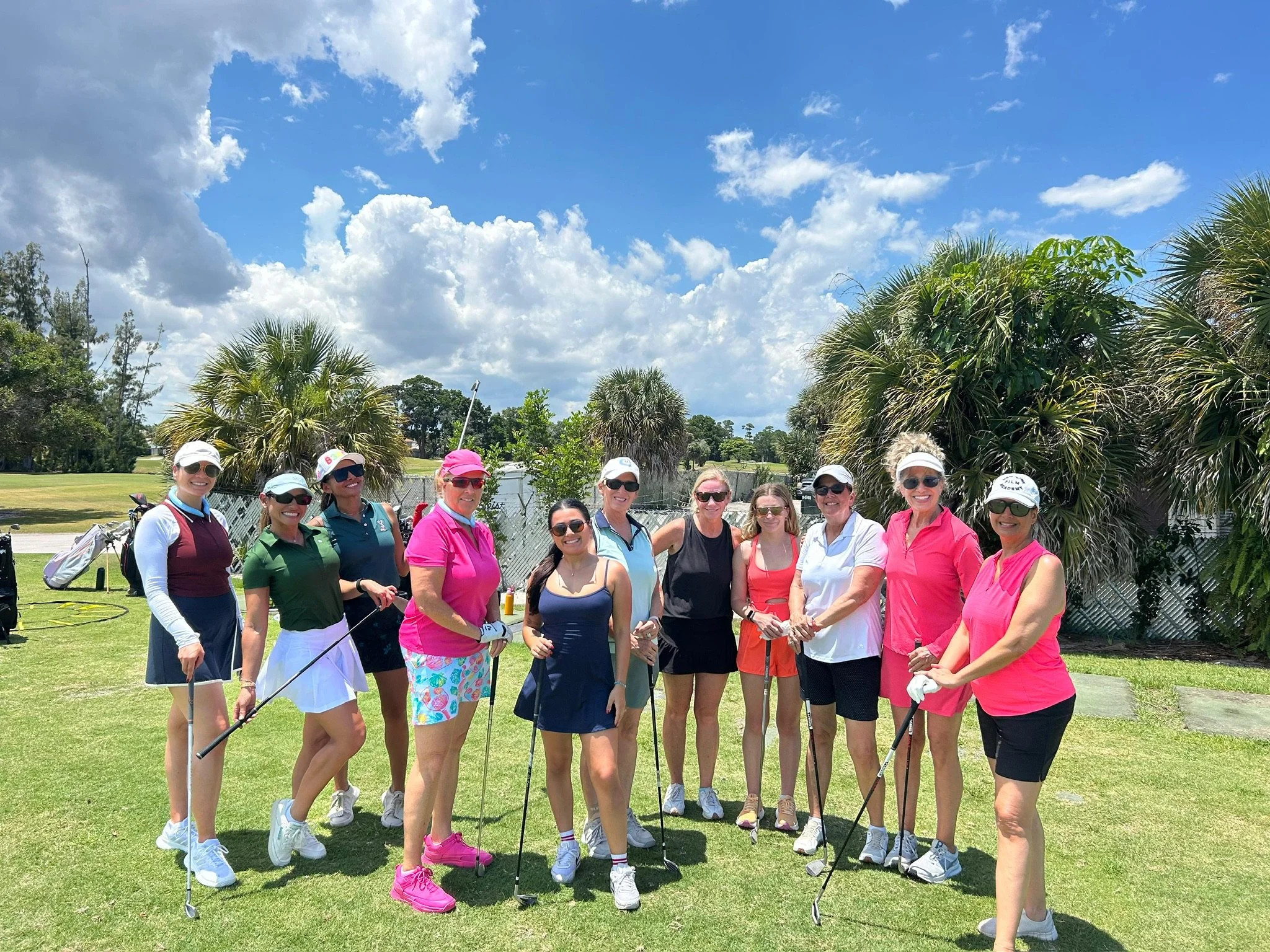 Group of women golfers posing on a golf course, dressed in colorful golf attire, with palm trees and a partly cloudy sky in the background.