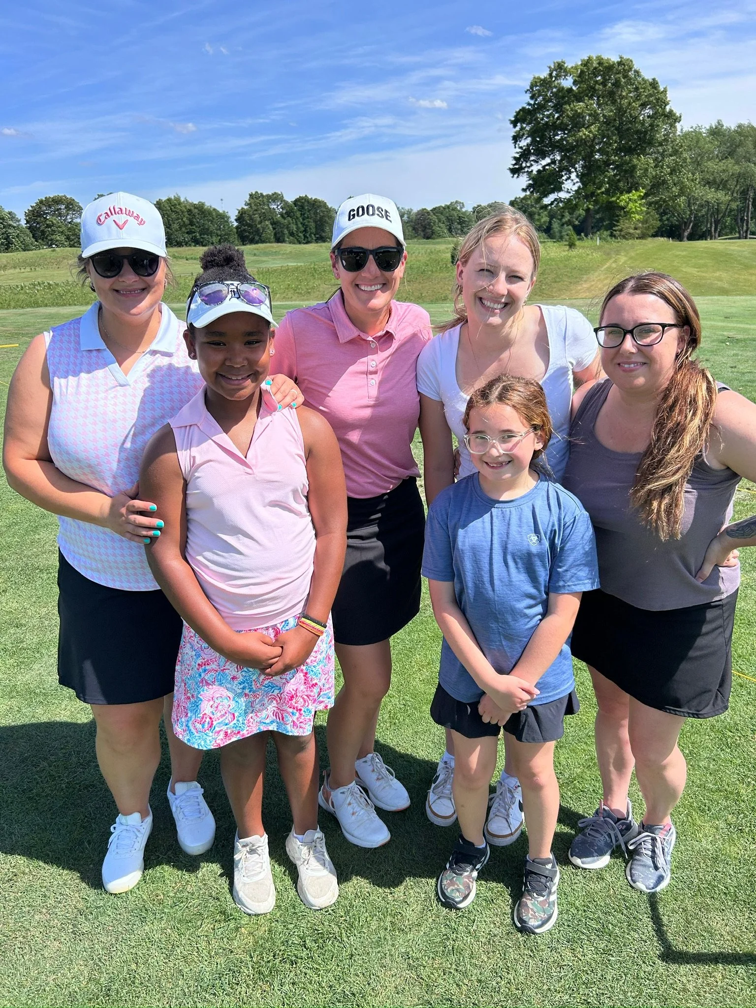 Group of seven women and girls on a golf course, smiling for a photo on a sunny day, wearing golf attire and hats, with green grass and trees in the background.