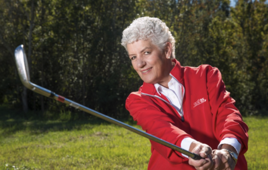 Older woman with short white hair wearing a red jacket, holding a golf club on a grassy course with trees in the background.