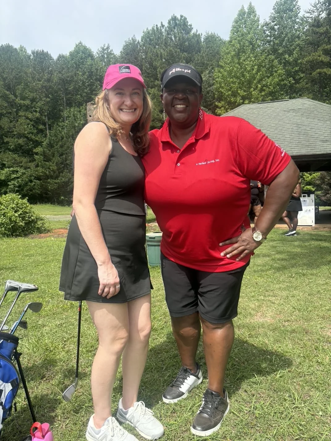Two women standing on a golf course, smiling for the camera. One woman is wearing a pink cap, black athletic dress, and white shoes. The other woman is wearing a black cap, red polo shirt, black shorts, and black sneakers. There is green grass, trees