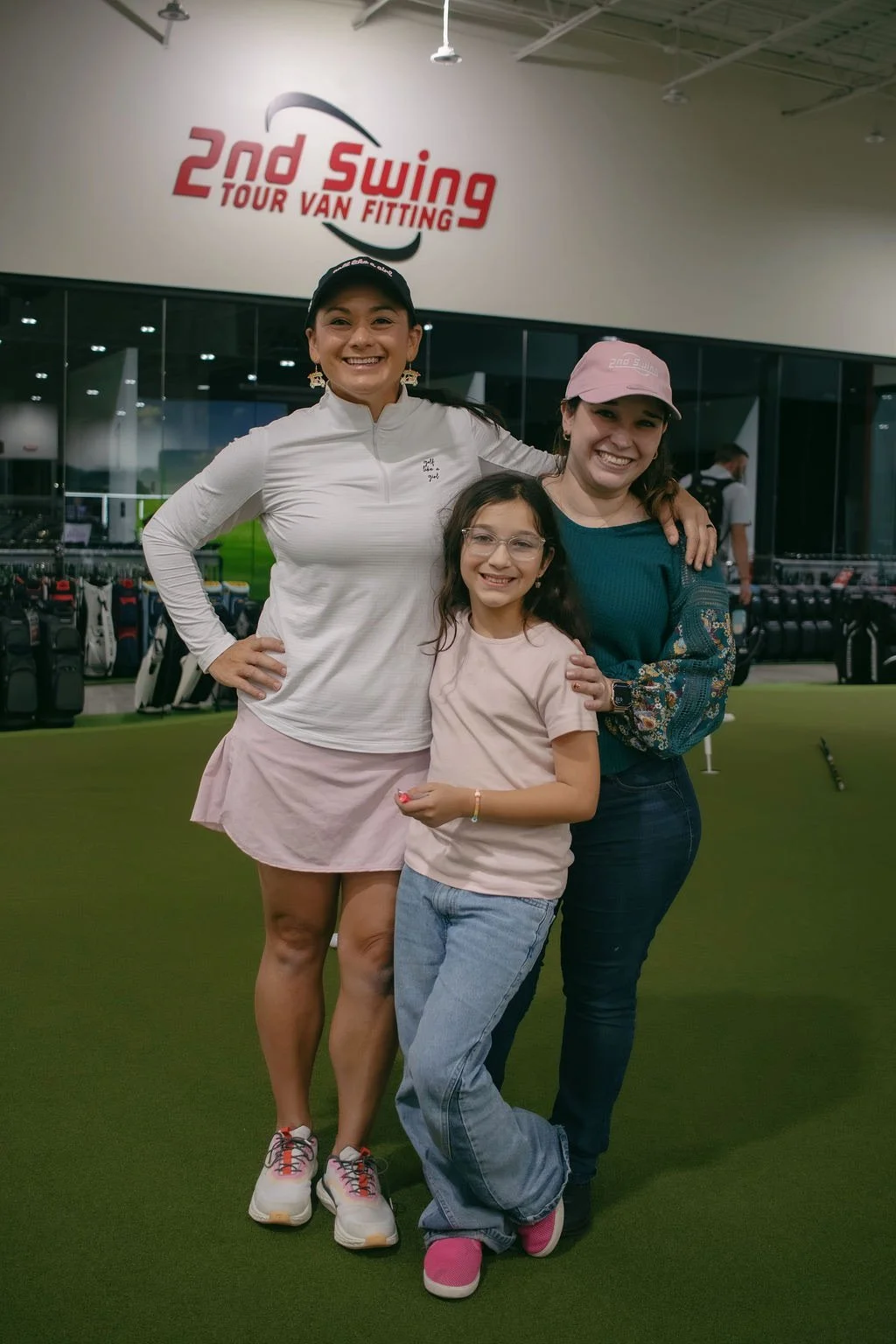 Three women and a girl smiling at a golf course indoors, with a sign that reads '2nd Swing Tour Van Fitting' in the background.