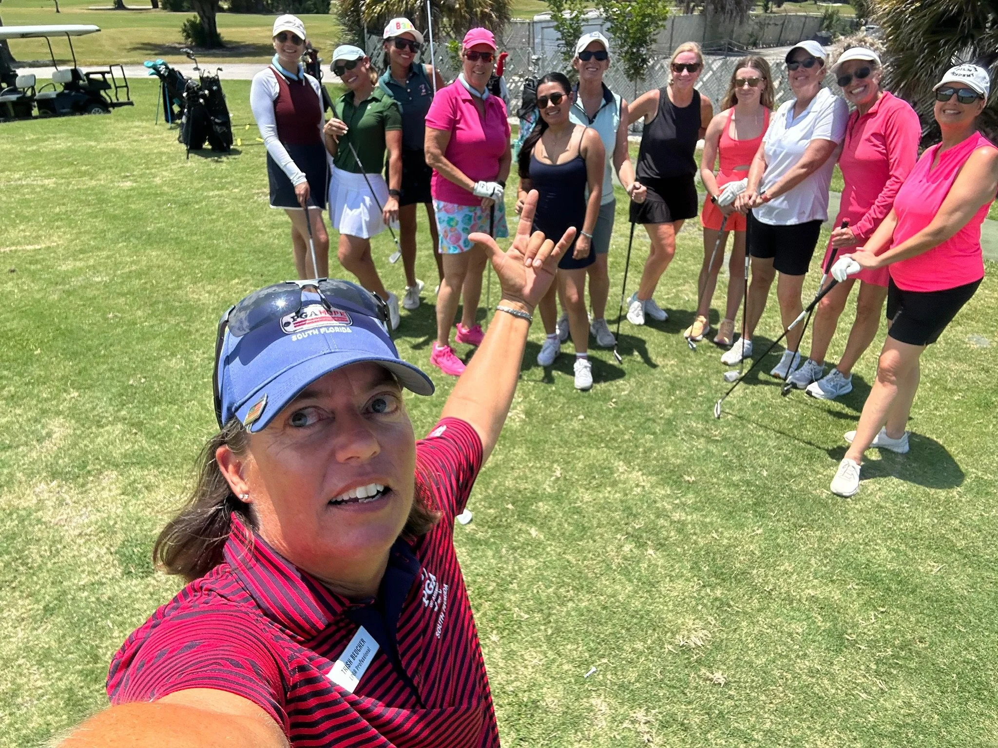 Group of women golfers standing on a golf course, smiling at the camera, with golf clubs and carts in the background.