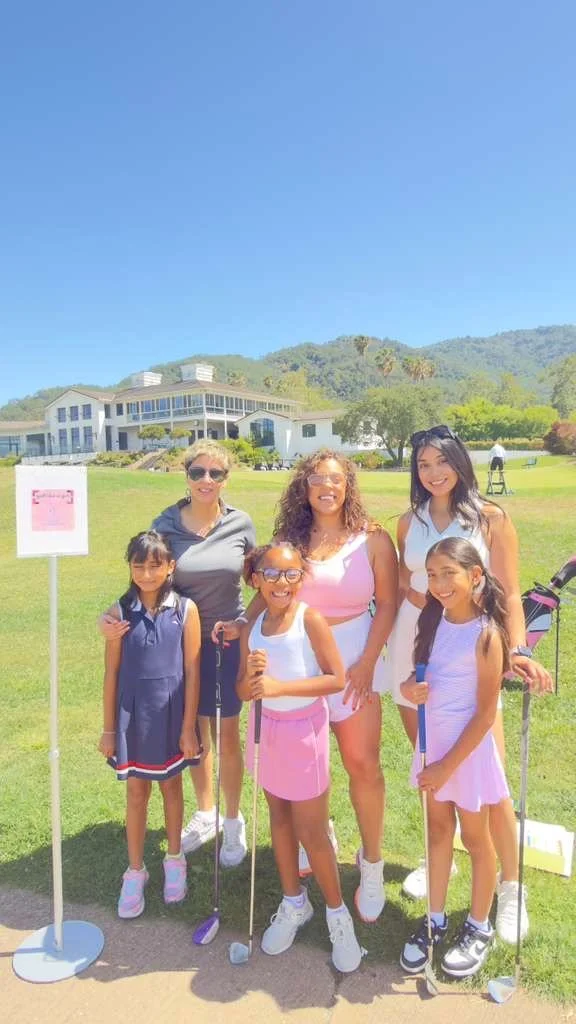 Group of six women and girls posing outdoors on a sunny day, holding golf clubs, with a building and hills in the background.