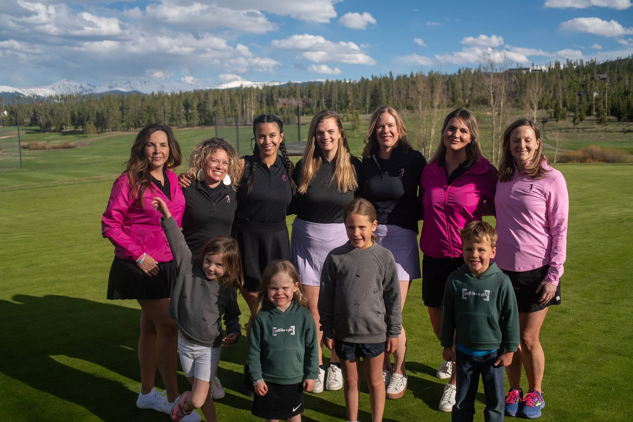 A group of women and children posing on a golf course with green grass, trees, and distant snow-capped mountains under a partly cloudy sky.