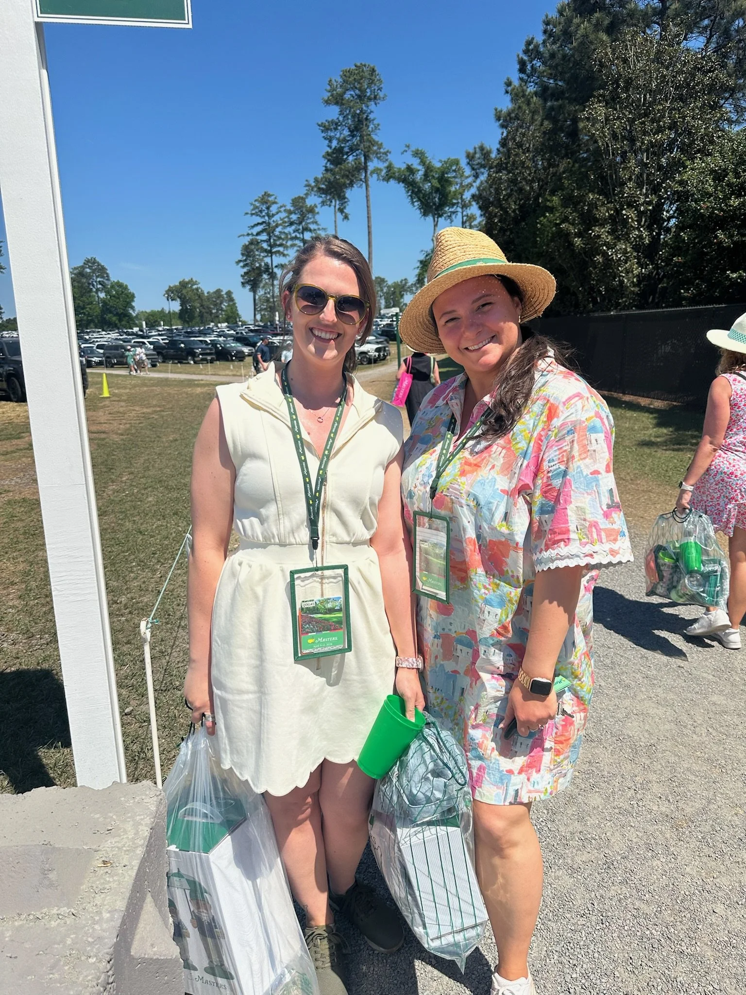Two women smiling outdoors on a sunny day, standing near a white frame with a parking lot and trees in the background. One woman has glasses and wears a white dress, the other has a straw hat and a colorful shirt. They are holding bags and wearing la