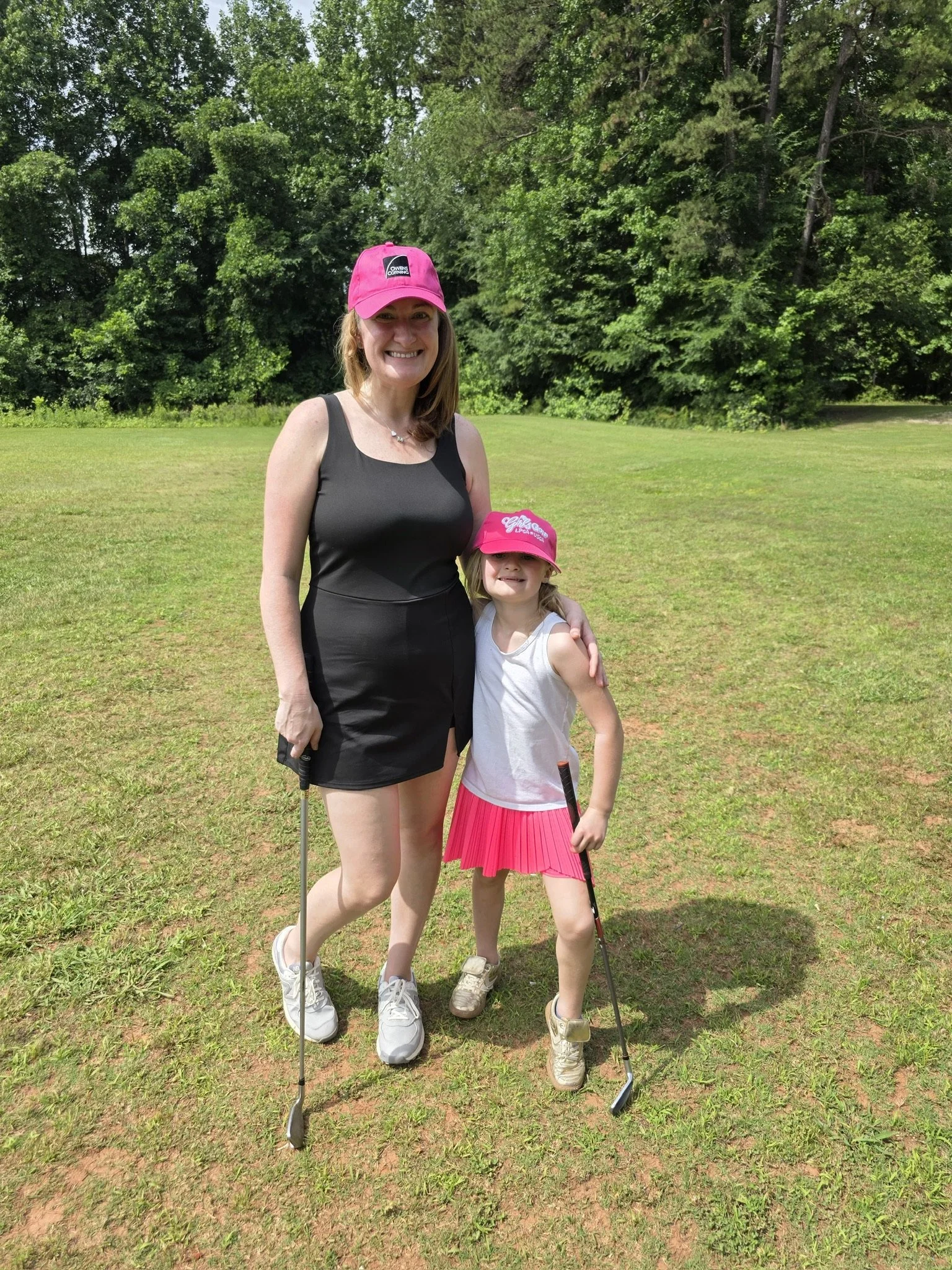 A woman and a young girl standing on a grassy area holding golf clubs outdoors, with green trees in the background. Both are smiling, and the girl is wearing a pink hat and pink skirt.