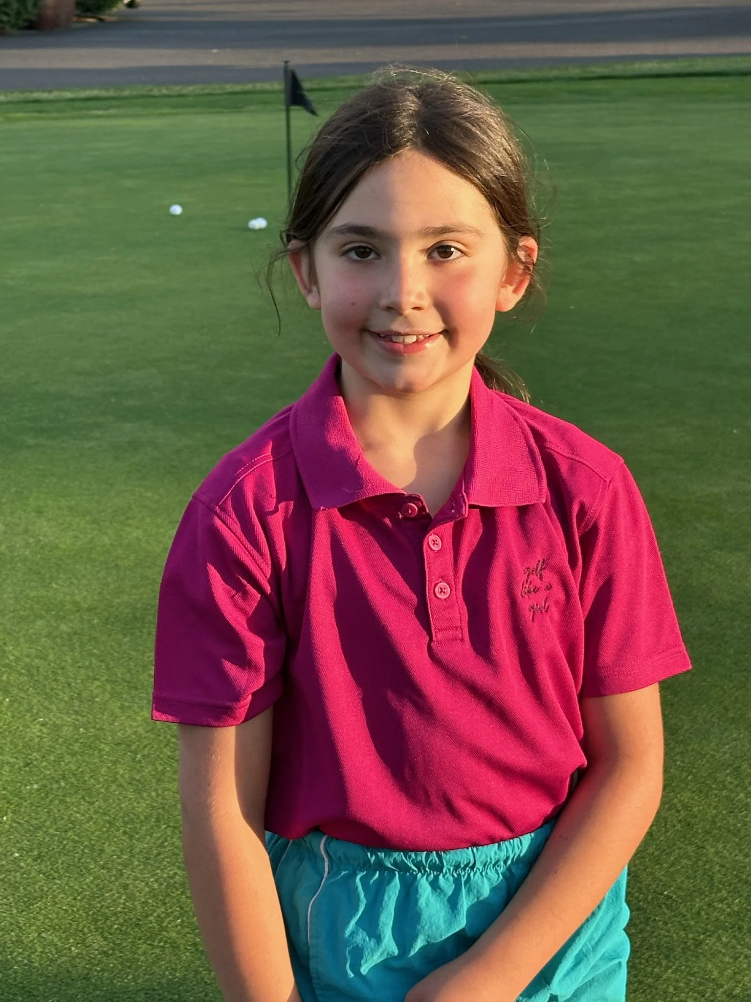 A young girl smiling, wearing a bright pink polo shirt and turquoise shorts, standing on a golf course with green grass and golf balls in the background.