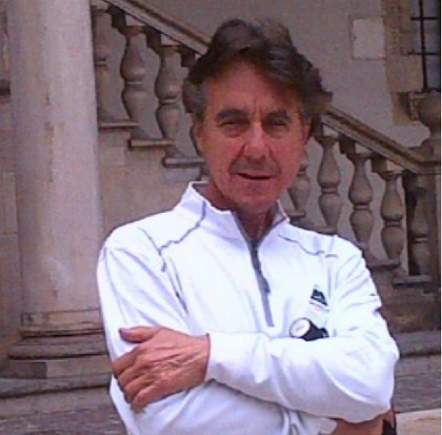 A man standing outdoors in front of a stone staircase and railing, wearing a white zip-up jacket with a watch on his wrist.