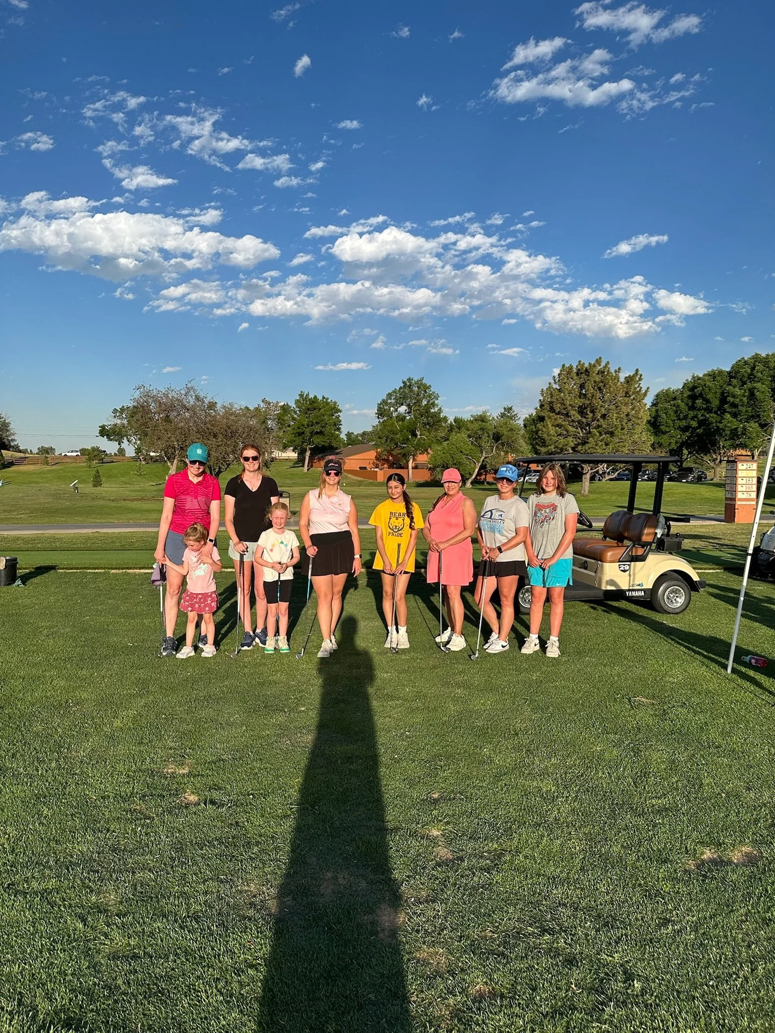 A group of nine people standing on a golf course with a golf cart behind them and a line of trees in the background, under a partly cloudy blue sky.